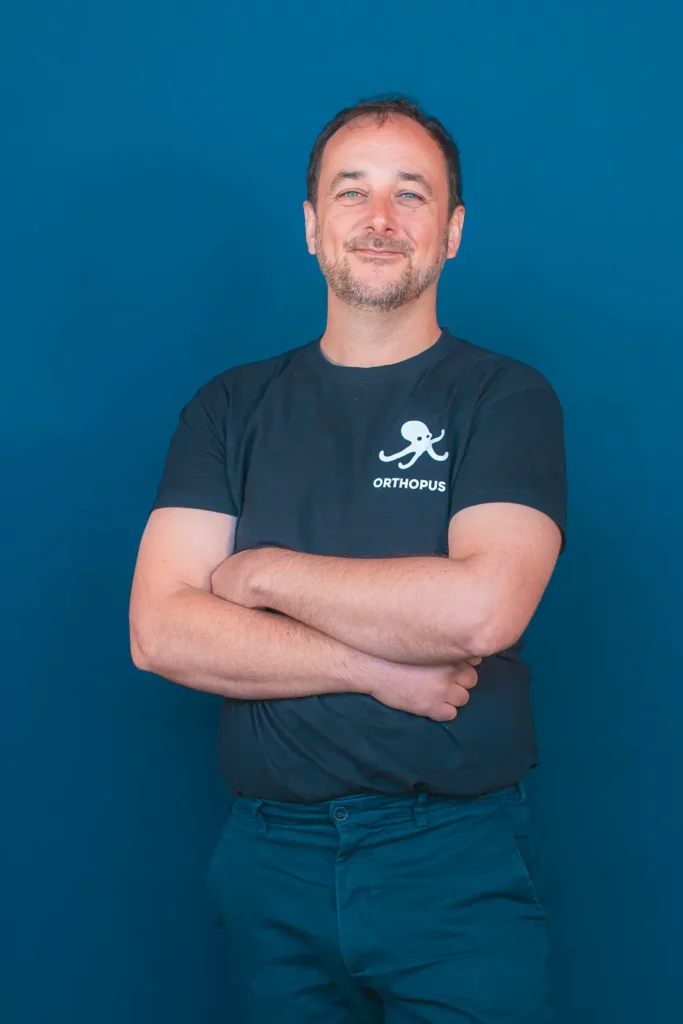 Portrait of a smiling man with arms crossed wearing a black T-shirt with an octopus logo, standing against a blue background.