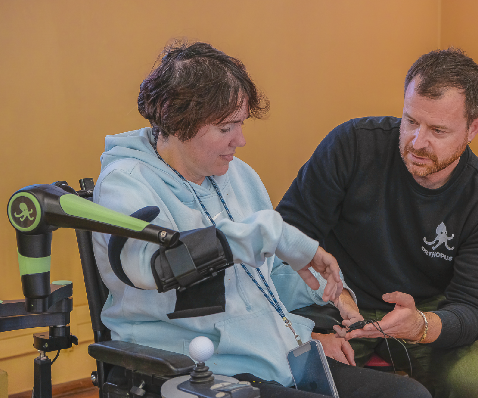 Person sitting in a powered wheelchair equipped with an arm support device, the ORTHOPUS Supporter, being assisted by another person who helps them use a small control. The scene takes place indoors, suggesting a rehabilitation or support setting.