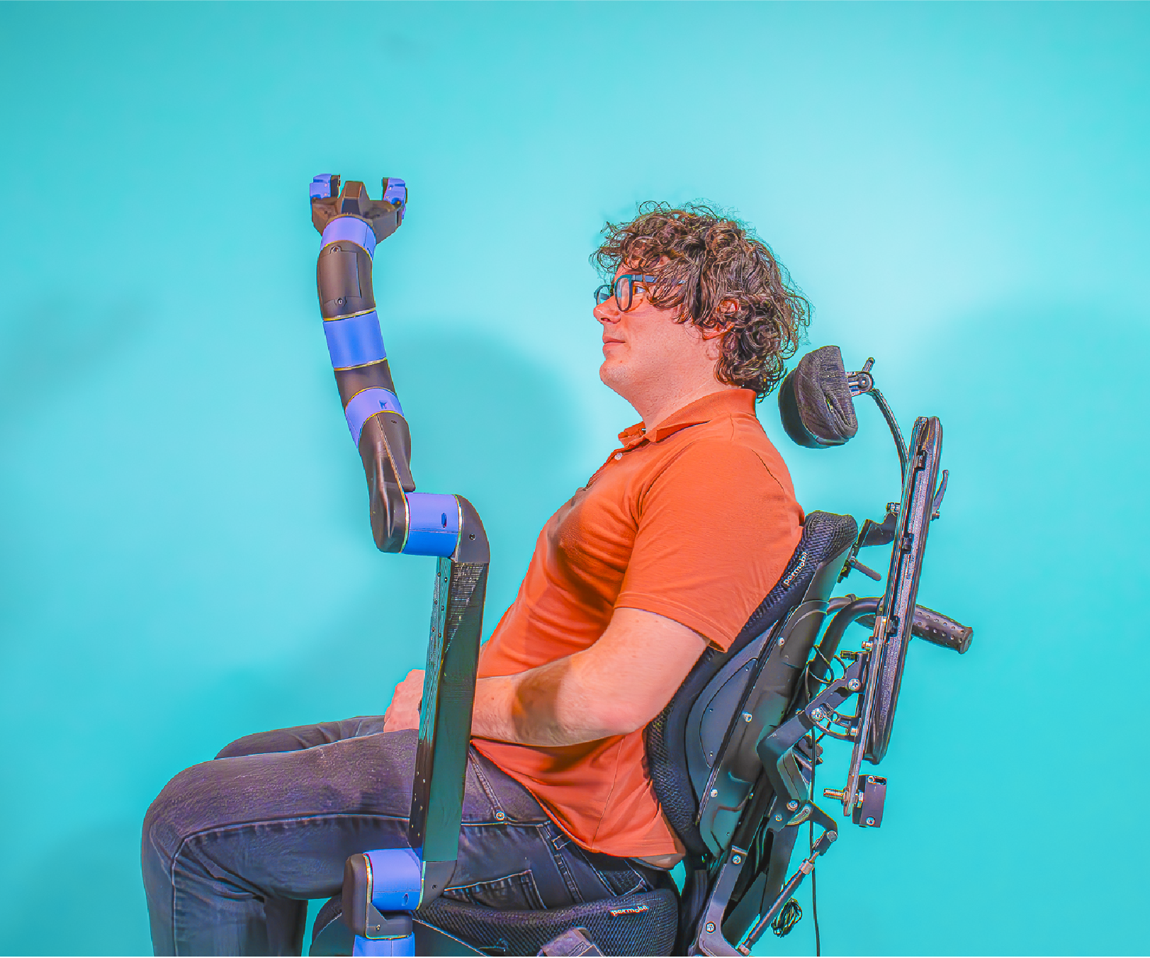 Side view of a man with curly hair and glasses sitting in a wheelchair against a turquoise background. A robotic arm support device is mounted to the wheelchair and positioned in front of him, aligned with his arm. He wears an orange shirt and jeans, with a headrest visible behind him.