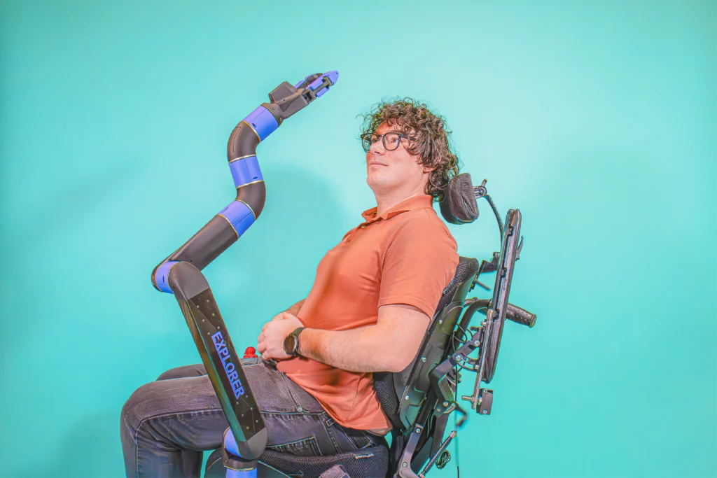 A curly-haired man wearing glasses and an orange polo shirt, seated in a powered wheelchair, equipped with a blue and black robotic assistive arm, the ORTHOPUS Explorer, extending upward.