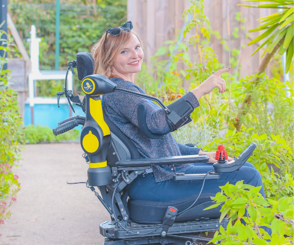 A smiling woman seated in a powered wheelchair outdoors in a garden, using the ORTHOPUS Partner, a robotic assistive device for arm mobility. The device supports her arm as she points toward plants, with greenery and a bright, natural setting in the background.