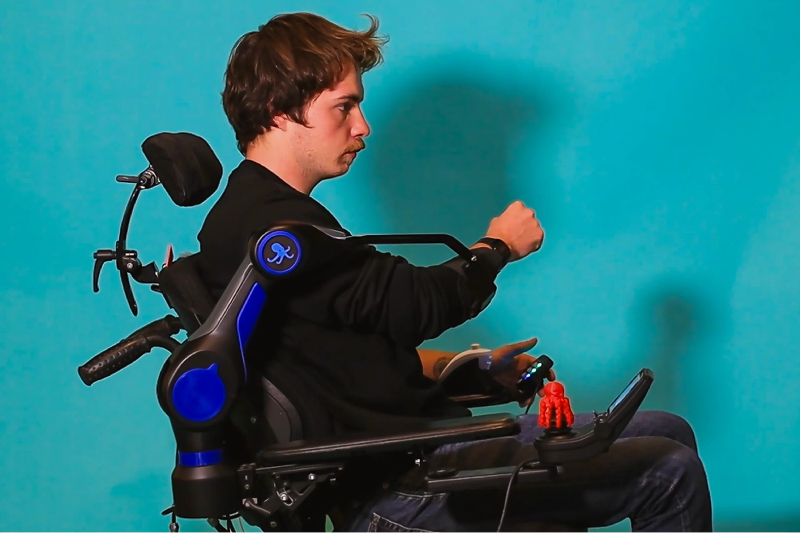 A man in a wheelchair wears a robotic assistive device, the ORTHOPUS Supporter, to aid arm mobility and poses in front of a blue background.