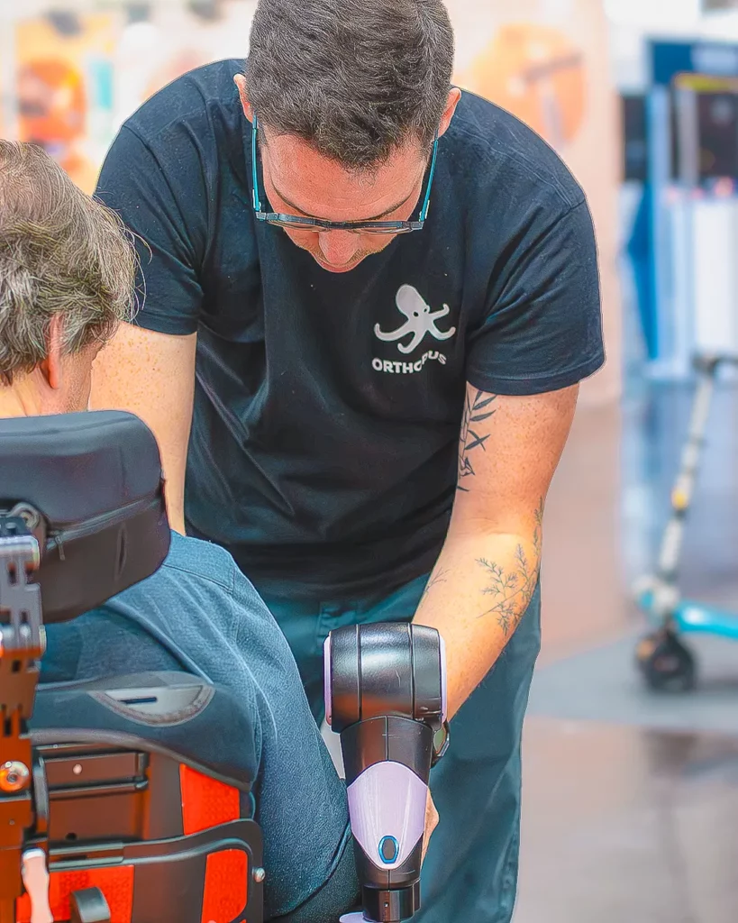 An ORTHOPUS engineer adjusting a robotic arm on the wheelchair of a seated user outdoors