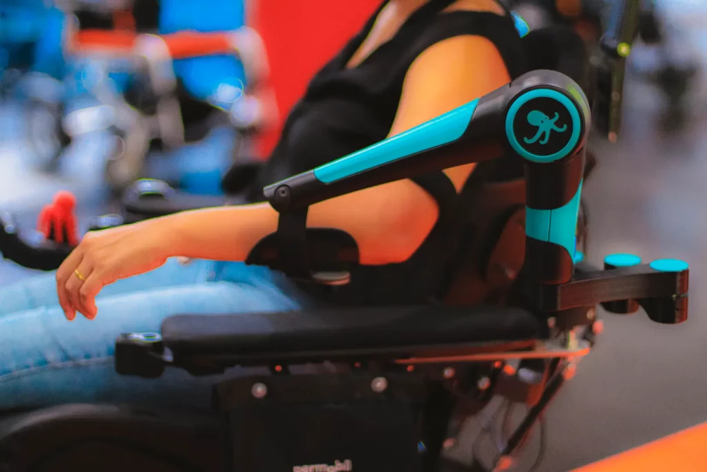 Close-up of the ORTHOPUS Supporter robotic arm in teal and black, mounted on a wheelchair and supporting the arm of a user at a trade show.