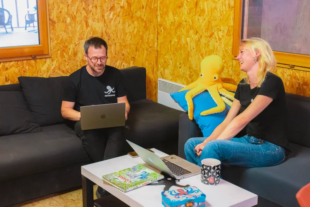 Two Orthopus team members sitting on a sofa, smiling and working on laptops in a warm wooden office space, with a large yellow octopus plush toy between them and Orthopus branded items on the coffee table.