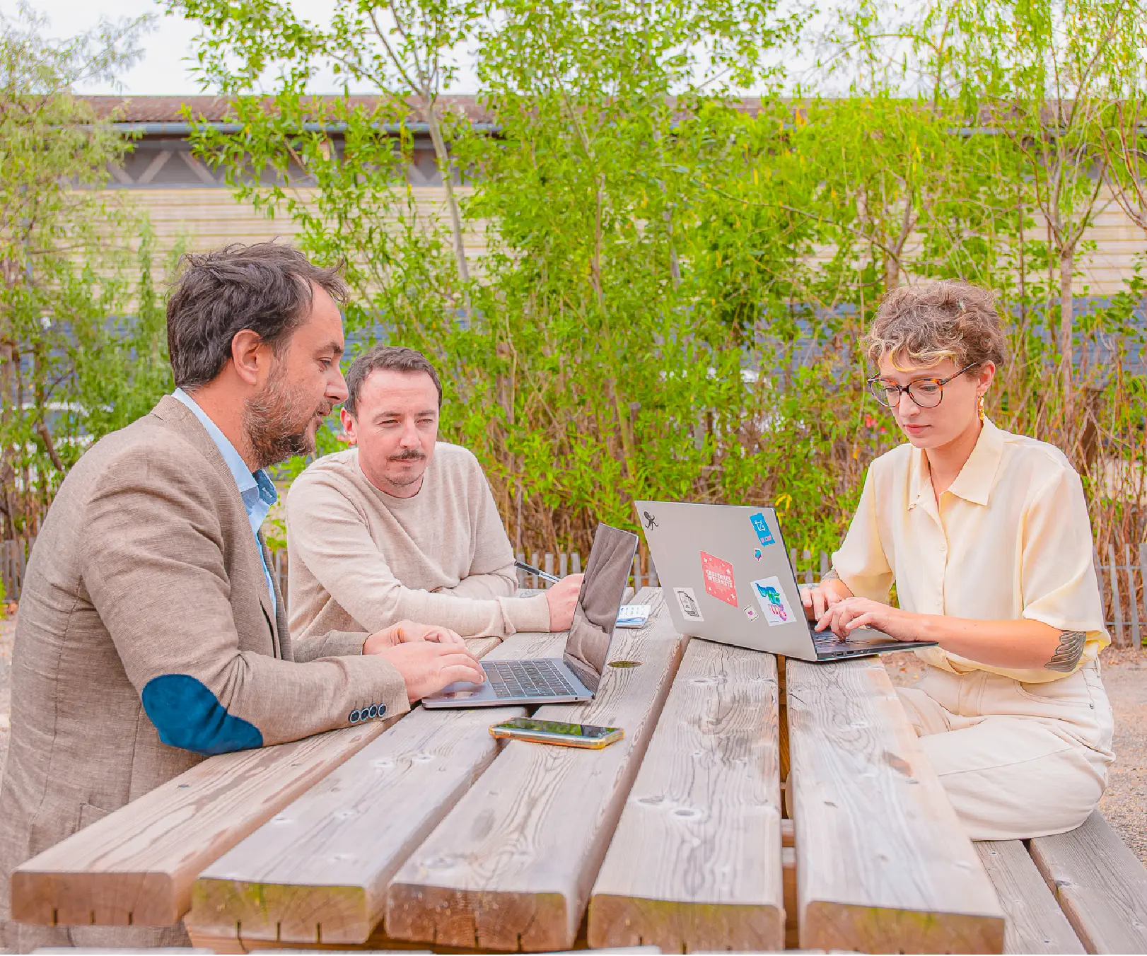 Three people working on laptops at a wooden table outdoors, surrounded by greenery.