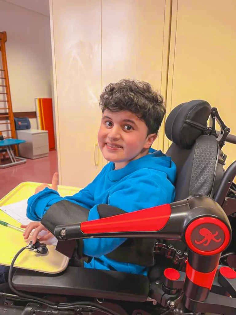 Young person sitting in a powered wheelchair at a desk, smiling and giving a thumbs up while using a robotic arm support and interact with a control device. Papers and a tray are placed in front of them, in an indoor classroom or therapy setting.