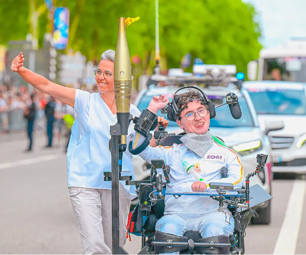 A smiling wheelchair user carrying the Paris 2024 Olympic torch during the relay, using the ORTHOPUS Supporter, a robotic assistive device for arm mobility. The torch is held upright beside them, while a companion walks alongside and waves to the crowd, with vehicles and spectators in the background.