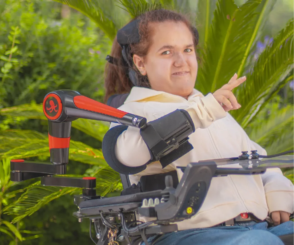 A person in a powered wheelchair using the ORTHOPUS Supporter, a robotic assistive device for arm mobility, outdoors in a green environment.