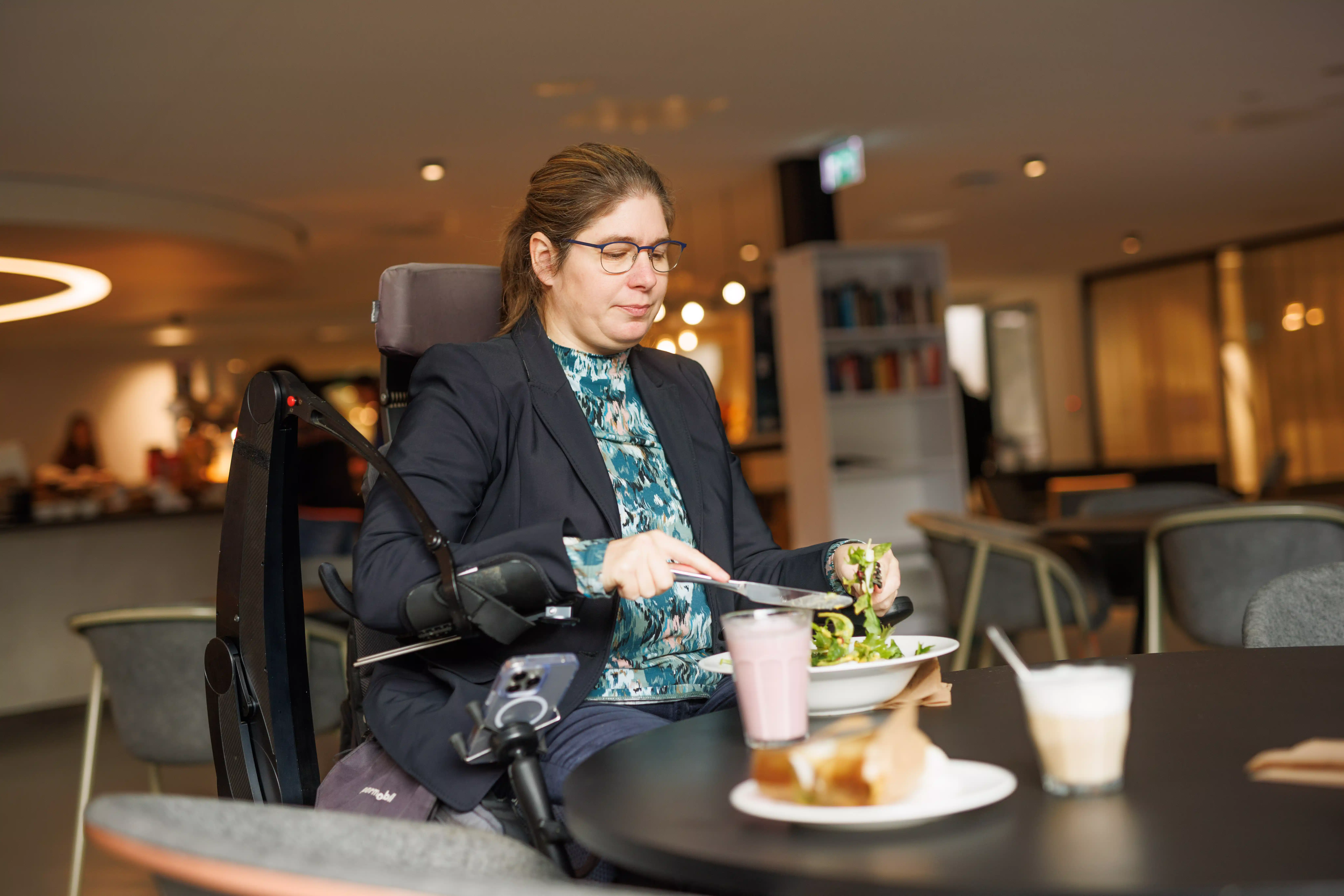 A woman in an electric wheelchair using a robotic arm to eat a meal independently at a restaurant table. 