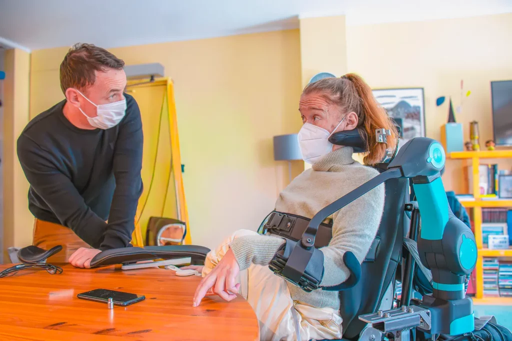 Person seated in a powered wheelchair equipped with a robotic arm (ORTHOPUS Partner) interacting with another person across a table during a beta test session.