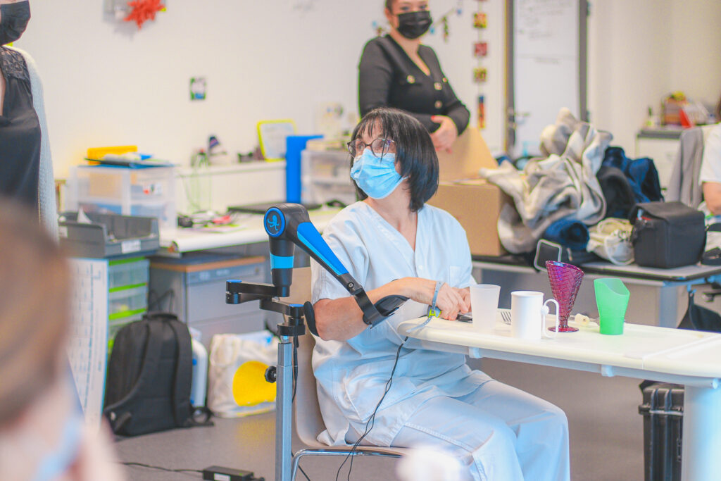 A healthcare professional in blue medical scrubs and a surgical mask, seated at a table, using a robotic assistive arm during a demonstration session, while a colleague watches in the background.