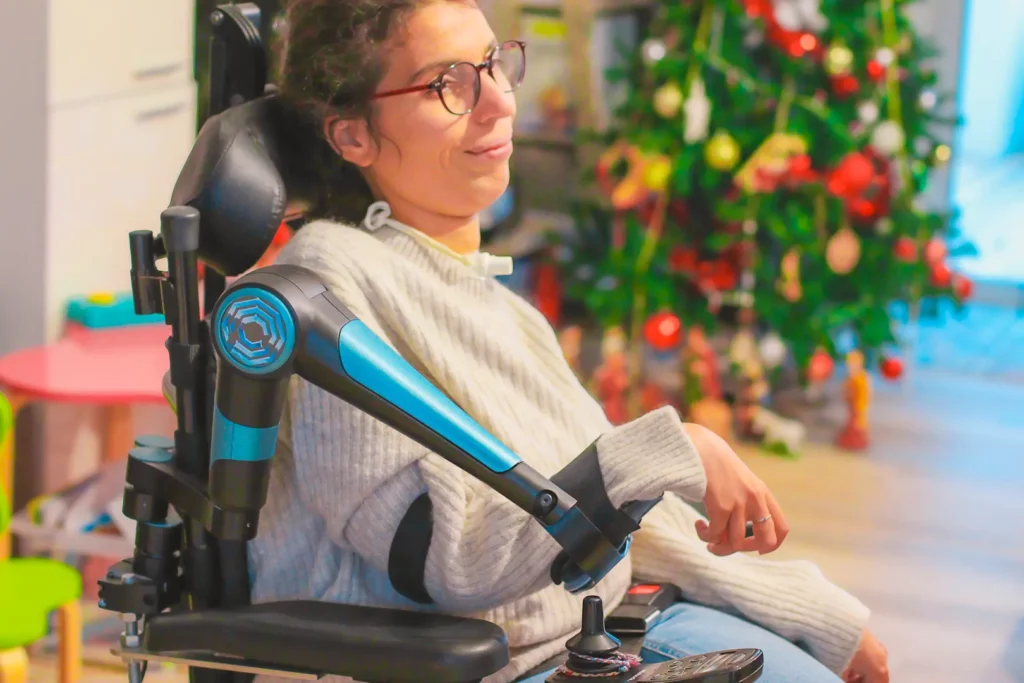 Femme souriante en fauteuil roulant électrique équipé d'un bras robotisé de soutien, devant un sapin de Noël décoré.